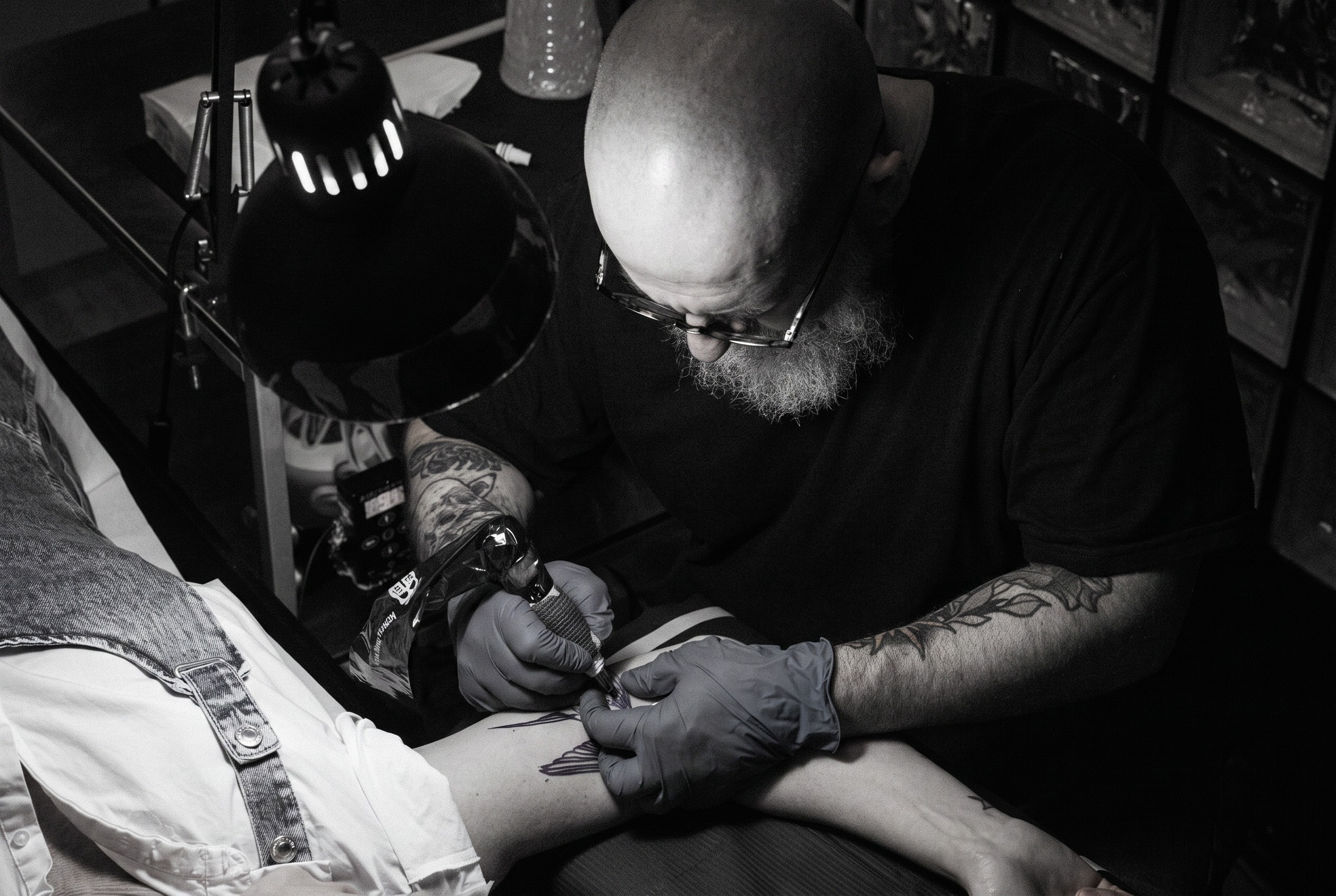 Tatoueur barbu penché sous une lampe d'atelier, machine en main, tatouant l'avant-bras d'un client